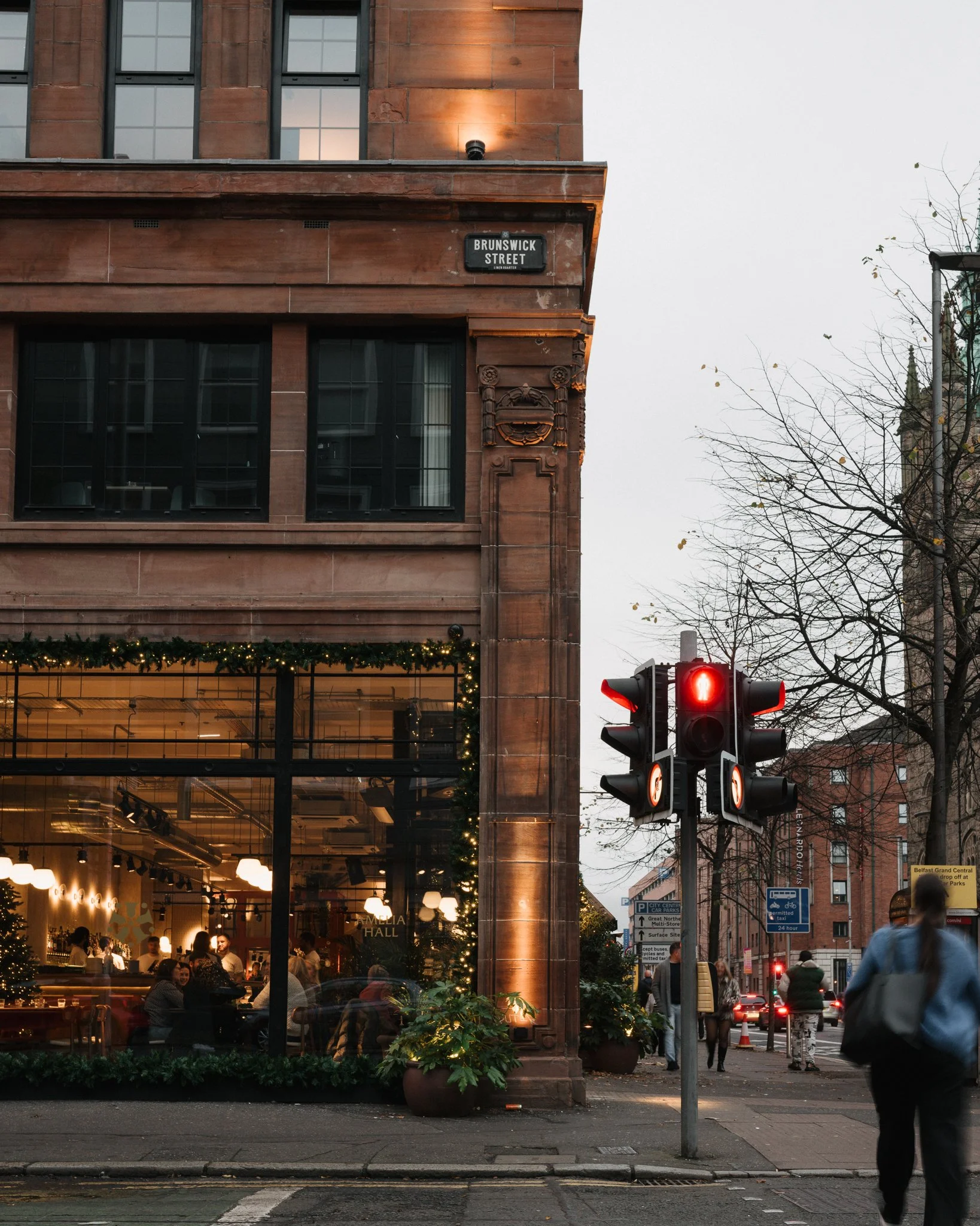Urban street scene showing a building with a sign that reads 'Brunswick Street,' a traffic light with a red signal, pedestrians crossing at a crosswalk, and a shop or café with large windows adorned with holiday lights, at dusk or early evening.