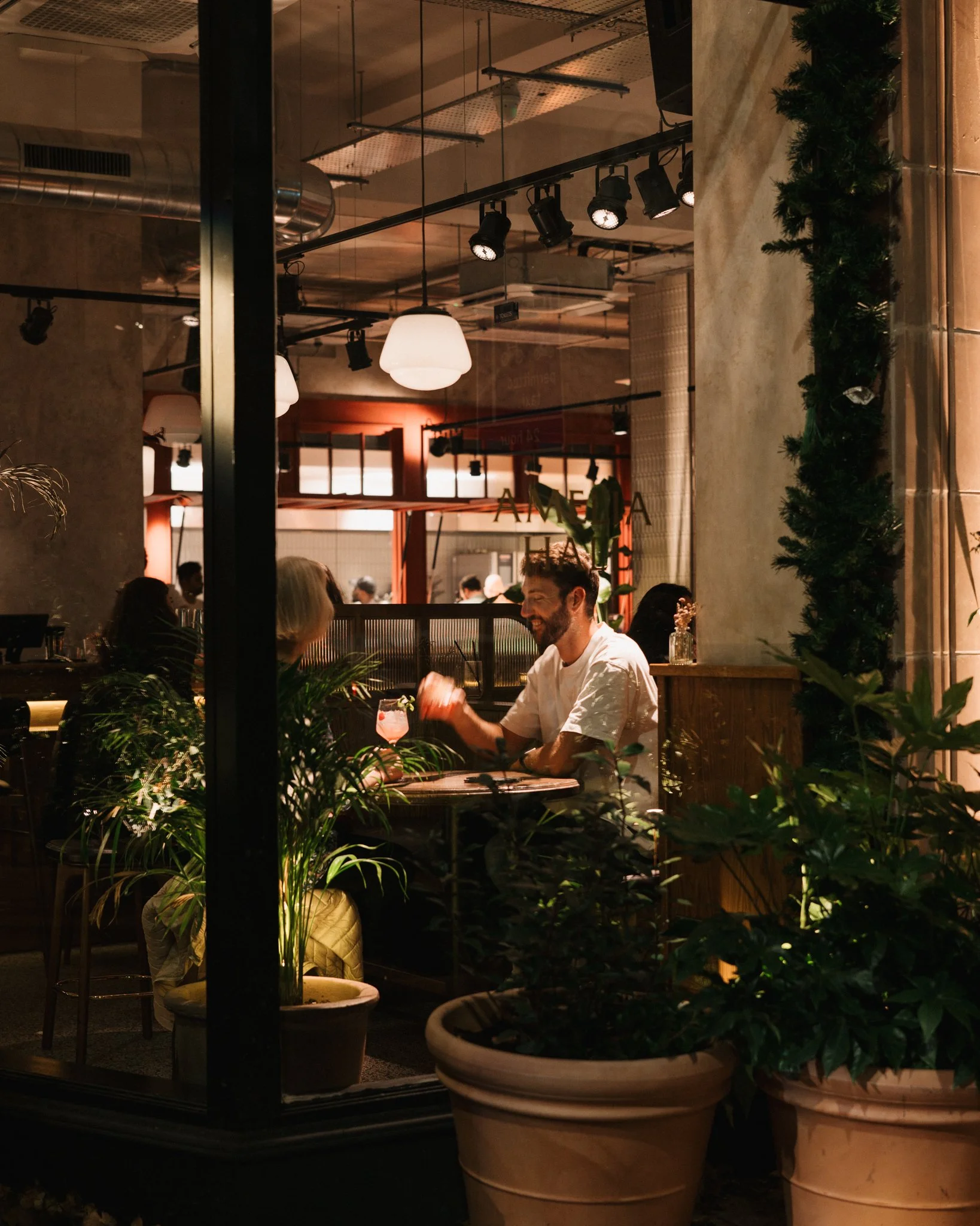 People dining in a restaurant seen through a glass window, with a man and woman smiling and talking. There are potted plants outside and modern ceiling lights inside.