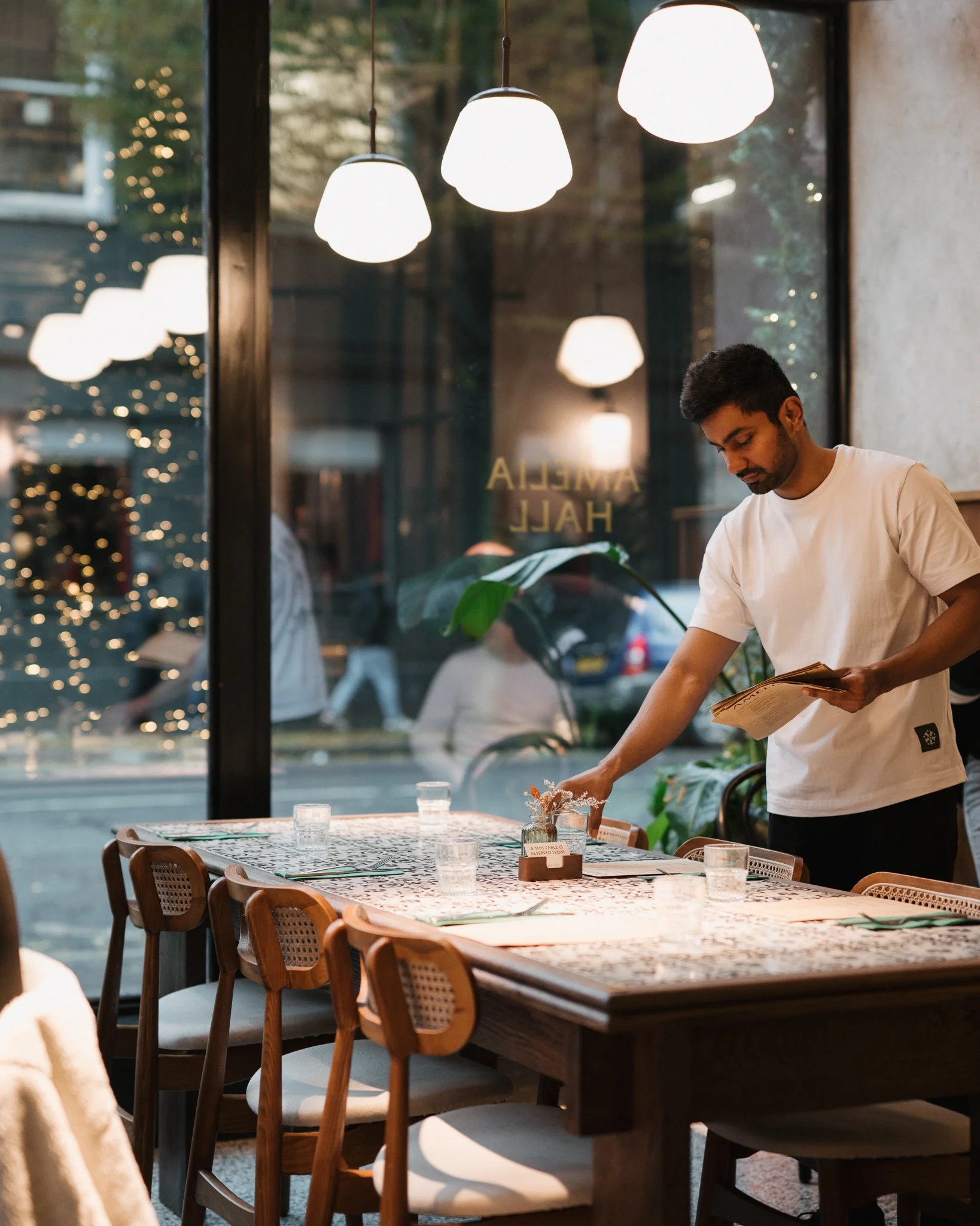 A man dressed in a white t-shirt arranging glassware on a dining table inside a restaurant with large glass windows, hanging white pendant lights, and city street view outside.