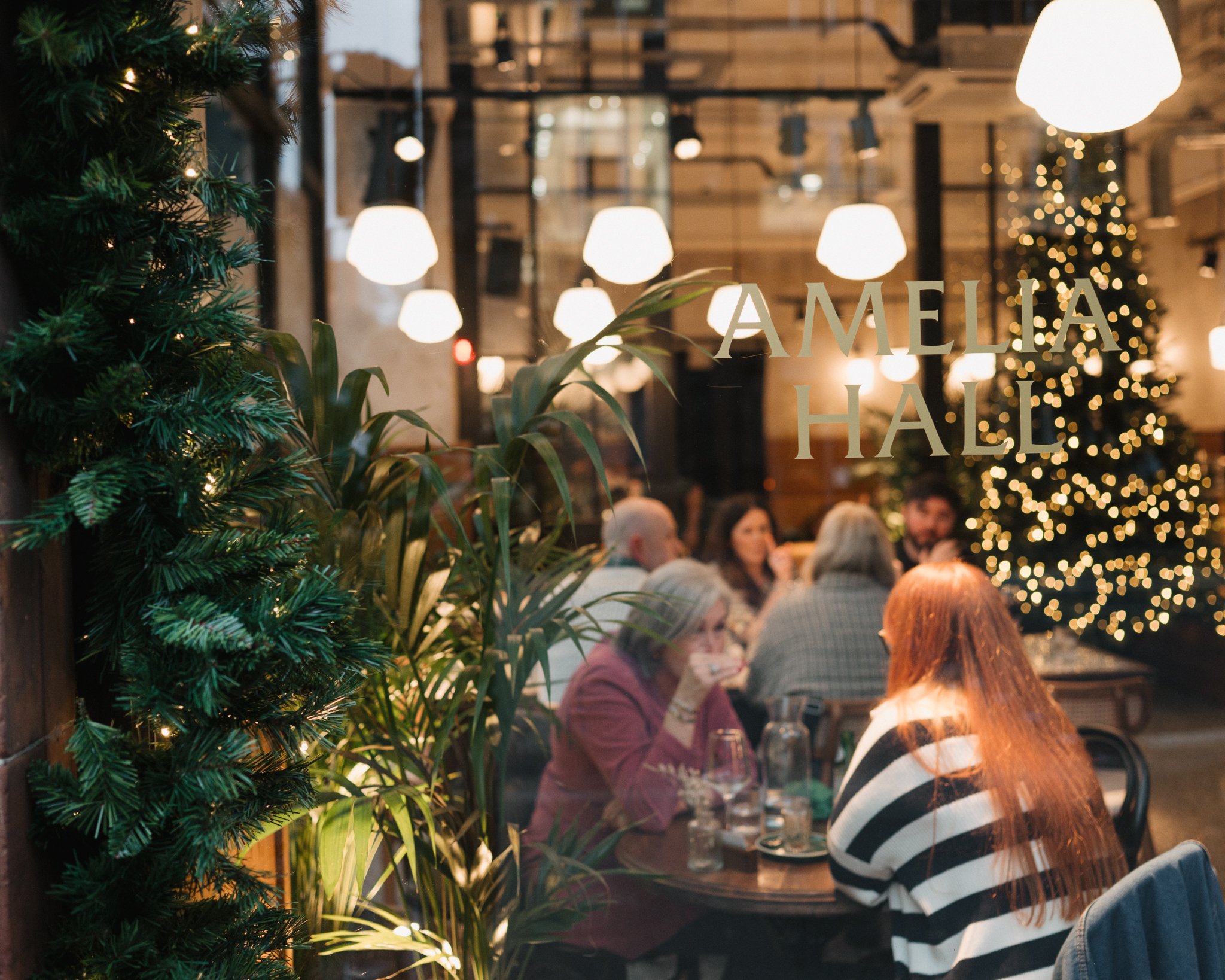 People dining at a restaurant with festive holiday decorations, including a Christmas tree and string lights, viewed through a glass window with "Amelia Hall" written on it.
