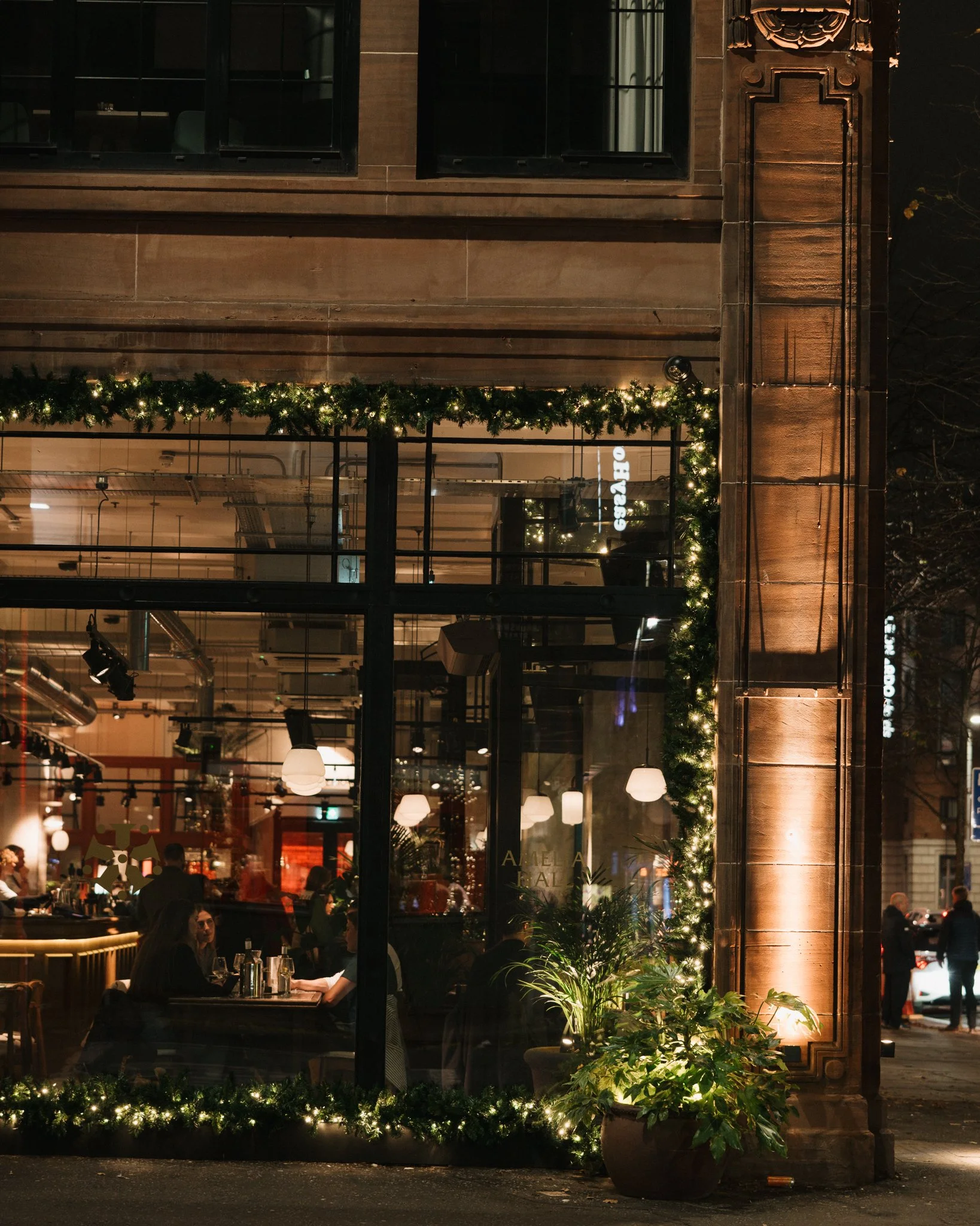 Nighttime view of a warmly lit restaurant with large glass windows decorated with Christmas garland and lights, showing people dining inside.