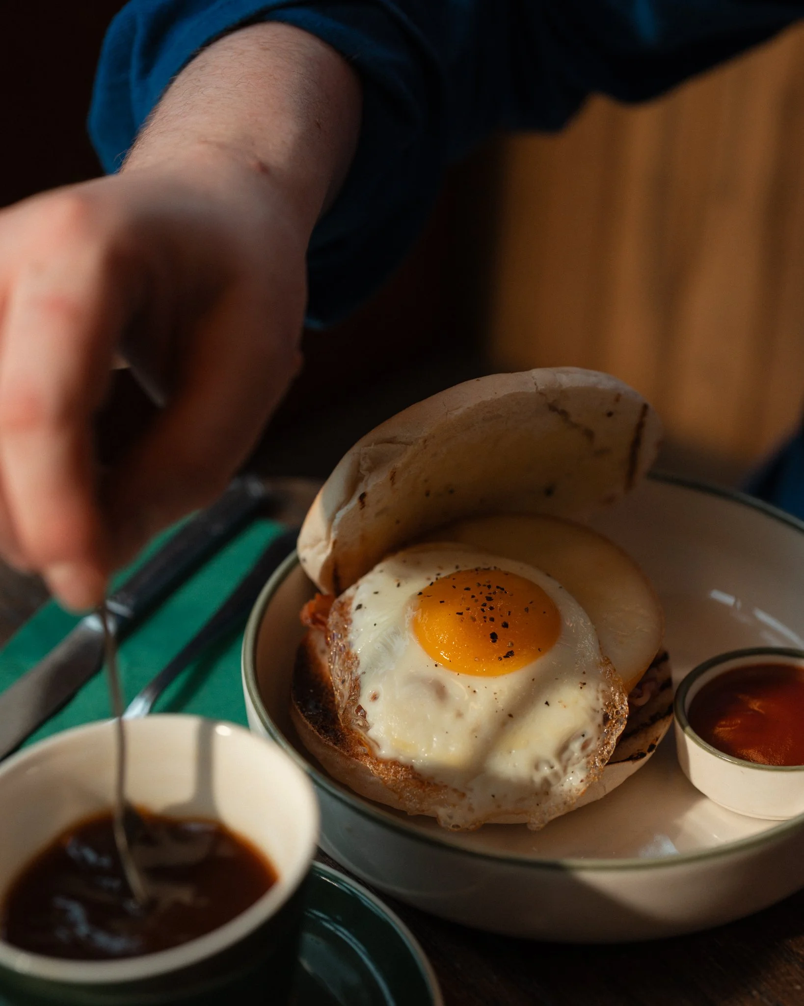 Close-up of a breakfast dish with a fried egg, toast, and a side of sauce, with a person's arm reaching over the table.