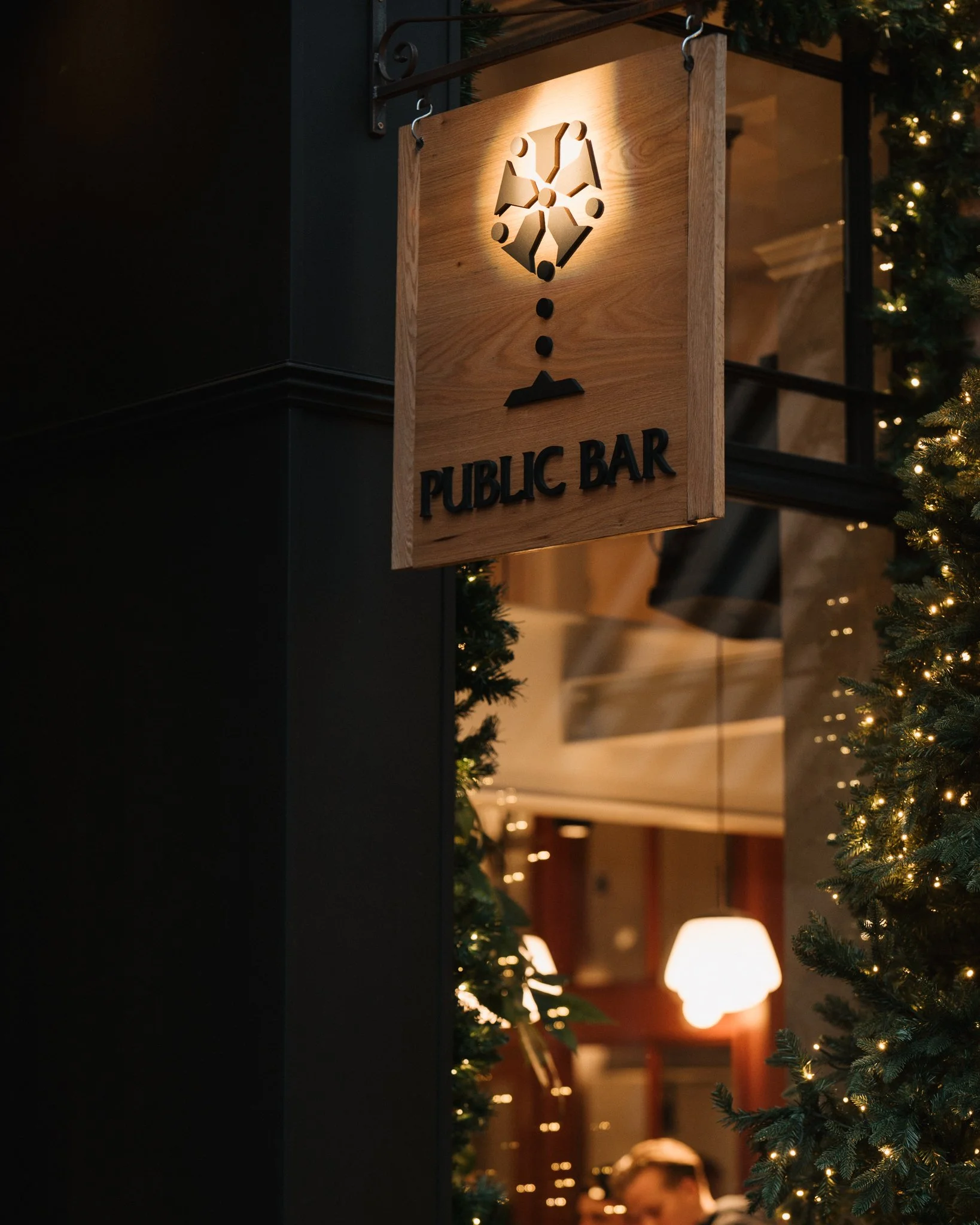 Wooden sign hanging at a public bar, illuminated, with a minimalist design of a dartboard and darts above the text 'Public Bar'. The background features Christmas trees and warm interior lighting.