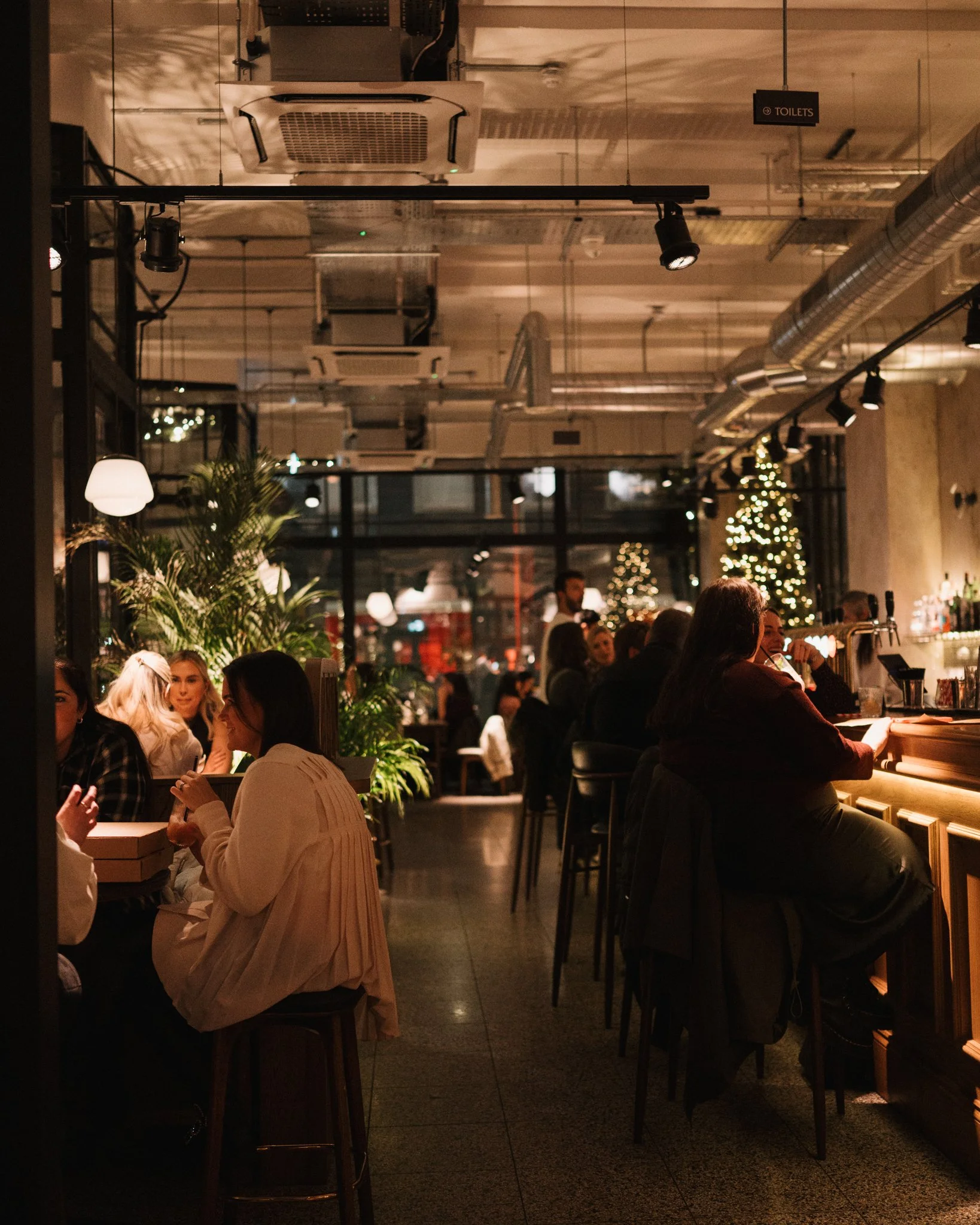 A cozy restaurant interior with warm lighting, people dining and socializing, decorated with Christmas trees and plants.