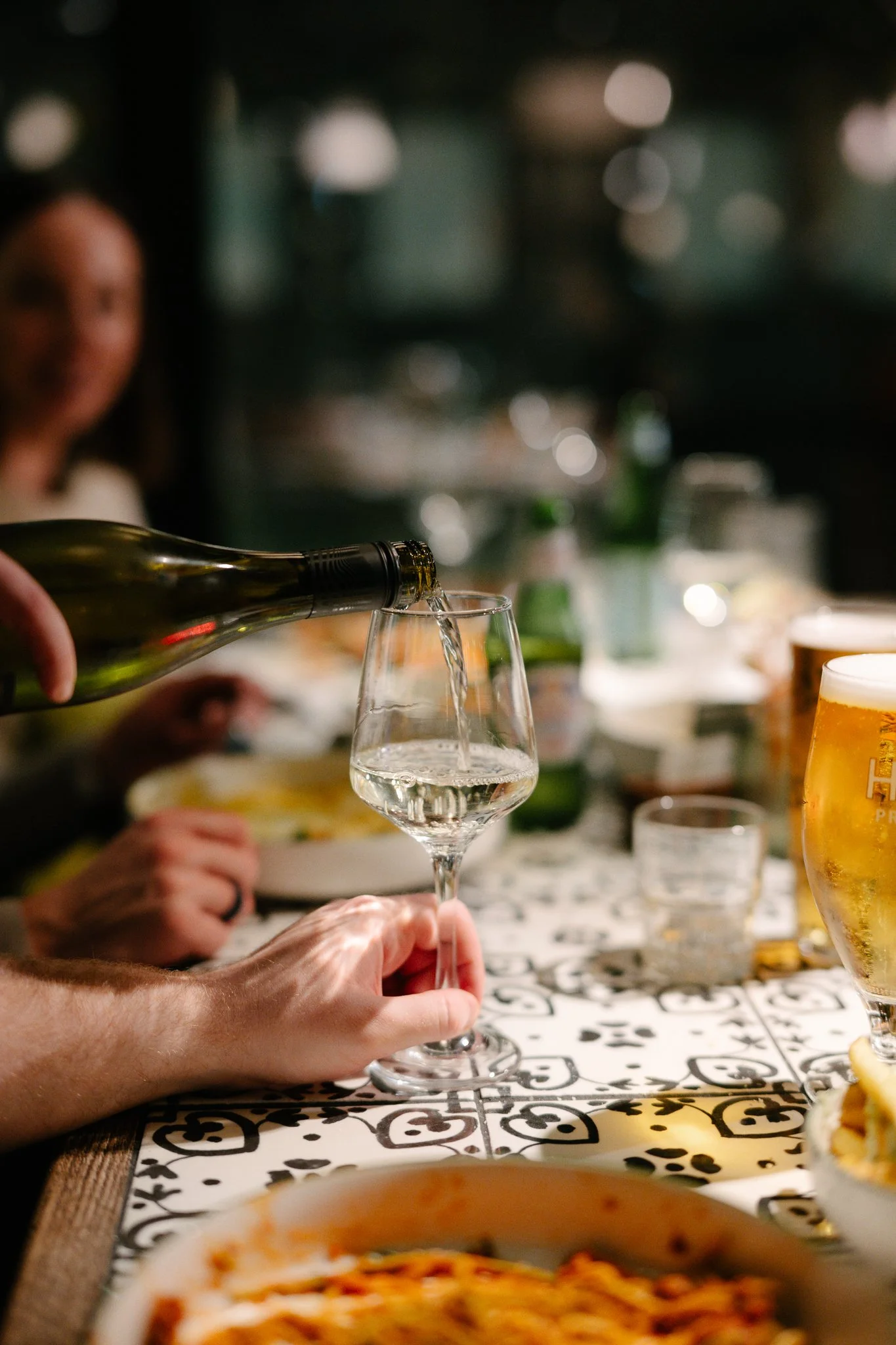 Person pouring white wine into a wine glass at a bar or restaurant table with food and drinks, including a beer glass, in the background.