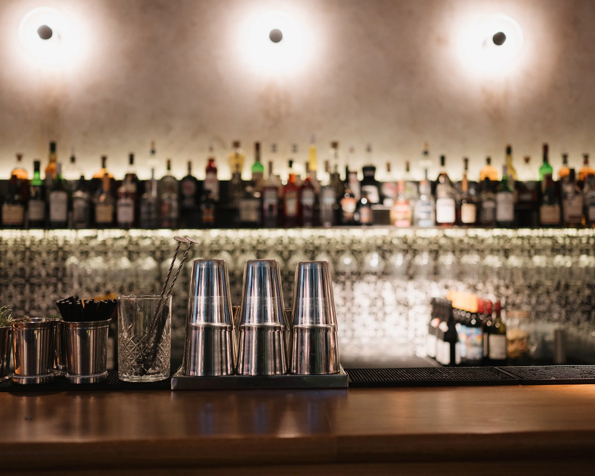 A bar counter with upside-down metal glasses, stir sticks in a textured glass, and small containers with bar tools, against a backdrop of a well-stocked bar with liquor bottles, illuminated by soft lighting.