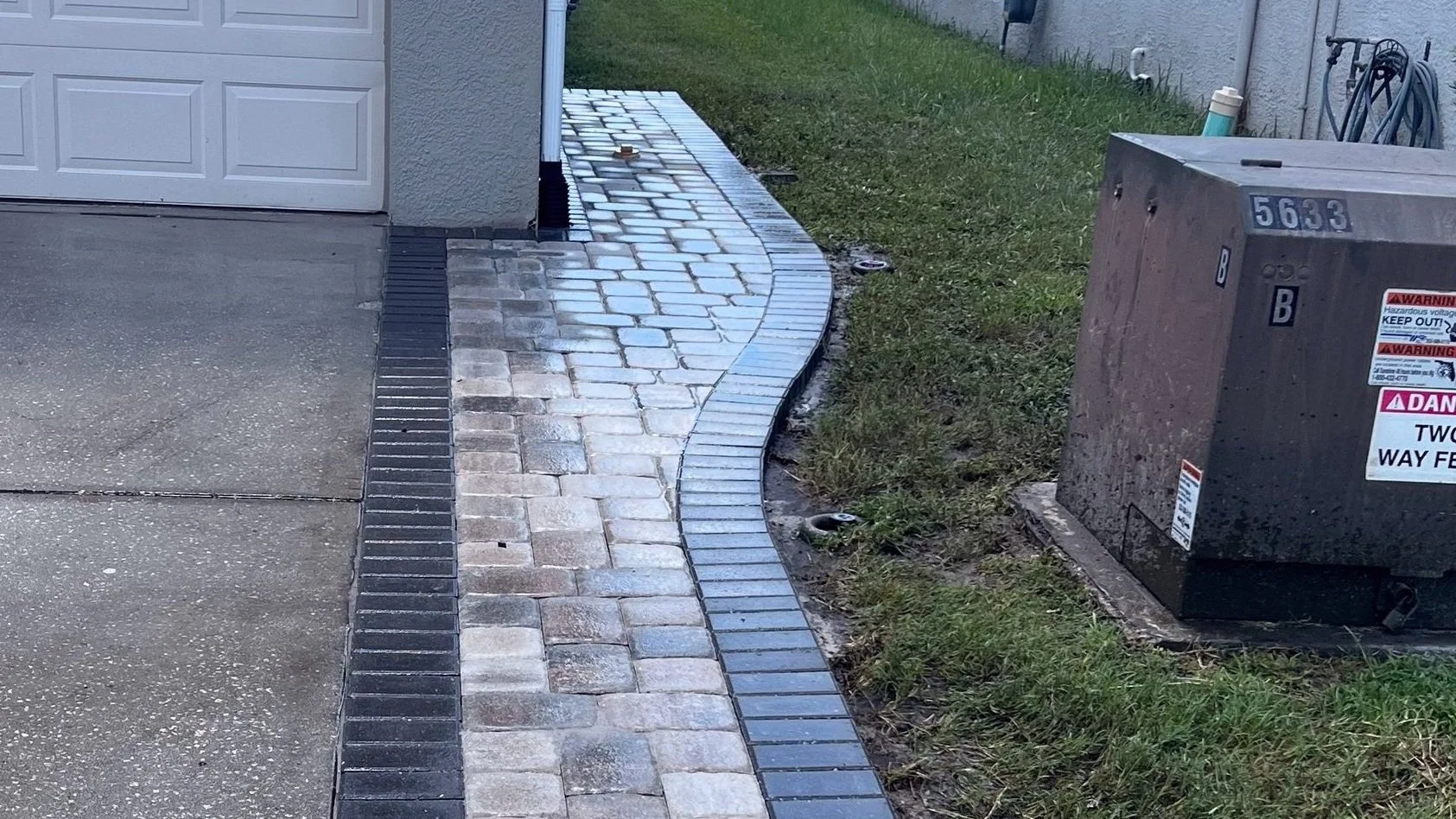 Curved gray paver walkway with a dark border beside a lawn and house