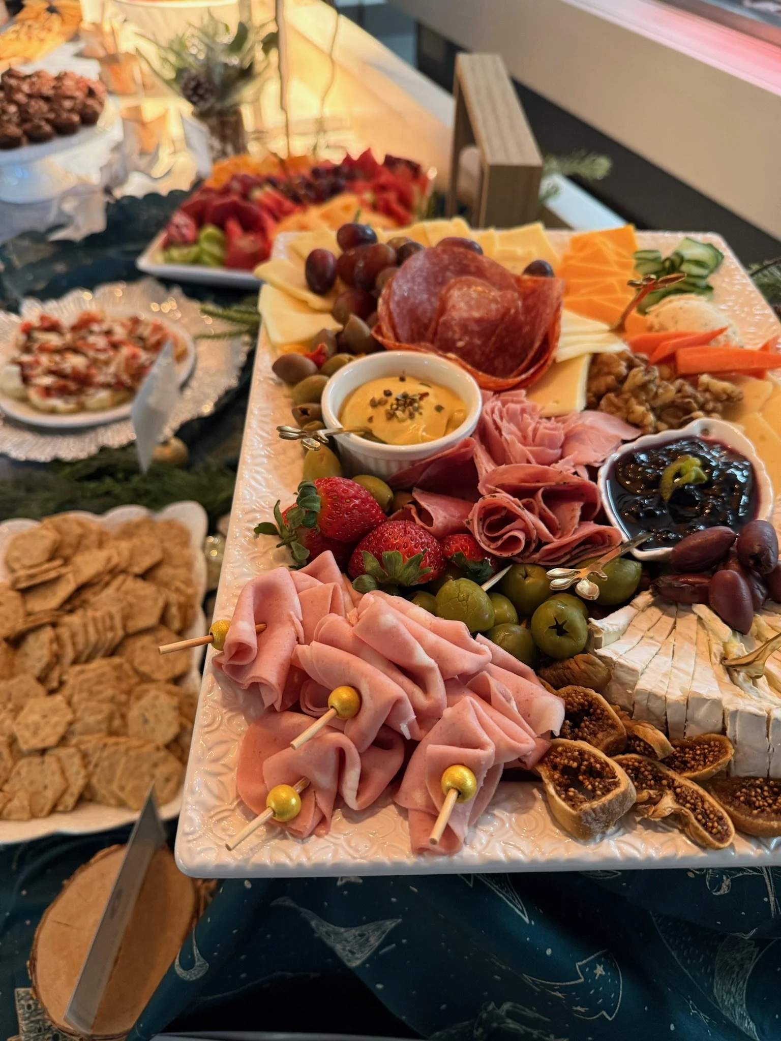 Charcuterie platter with cheeses, meats, strawberries, grapes, olives, fig slices, and dips, with additional finger foods and fruit arrangement in the background.