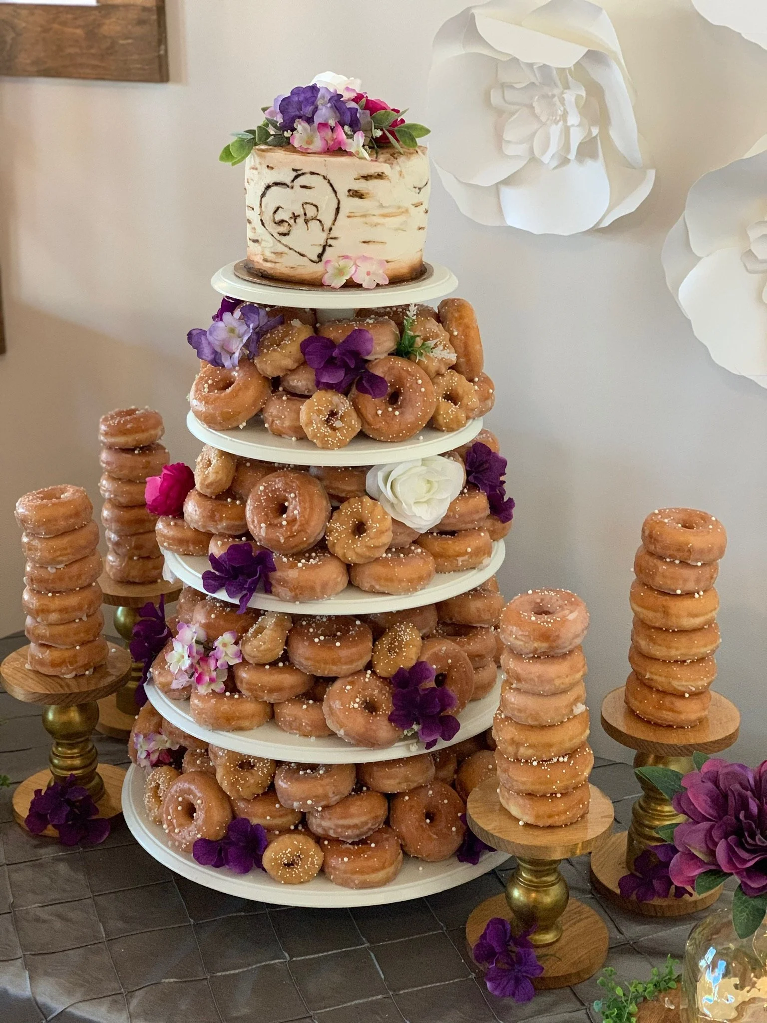 Donut tower with multiple tiers decorated with flowers, topped by a small cake with flowers and a heart drawing, and surrounded by more decorated stacks of donuts on wooden stands.