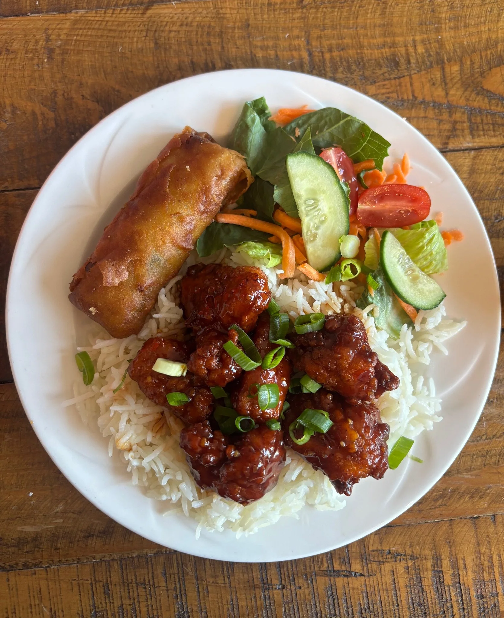 A plate of Chinese takeout food with spring rolls, chicken in sauce, white rice, and a side salad with cucumbers, tomatoes, carrots, and lettuce, on a wooden table.