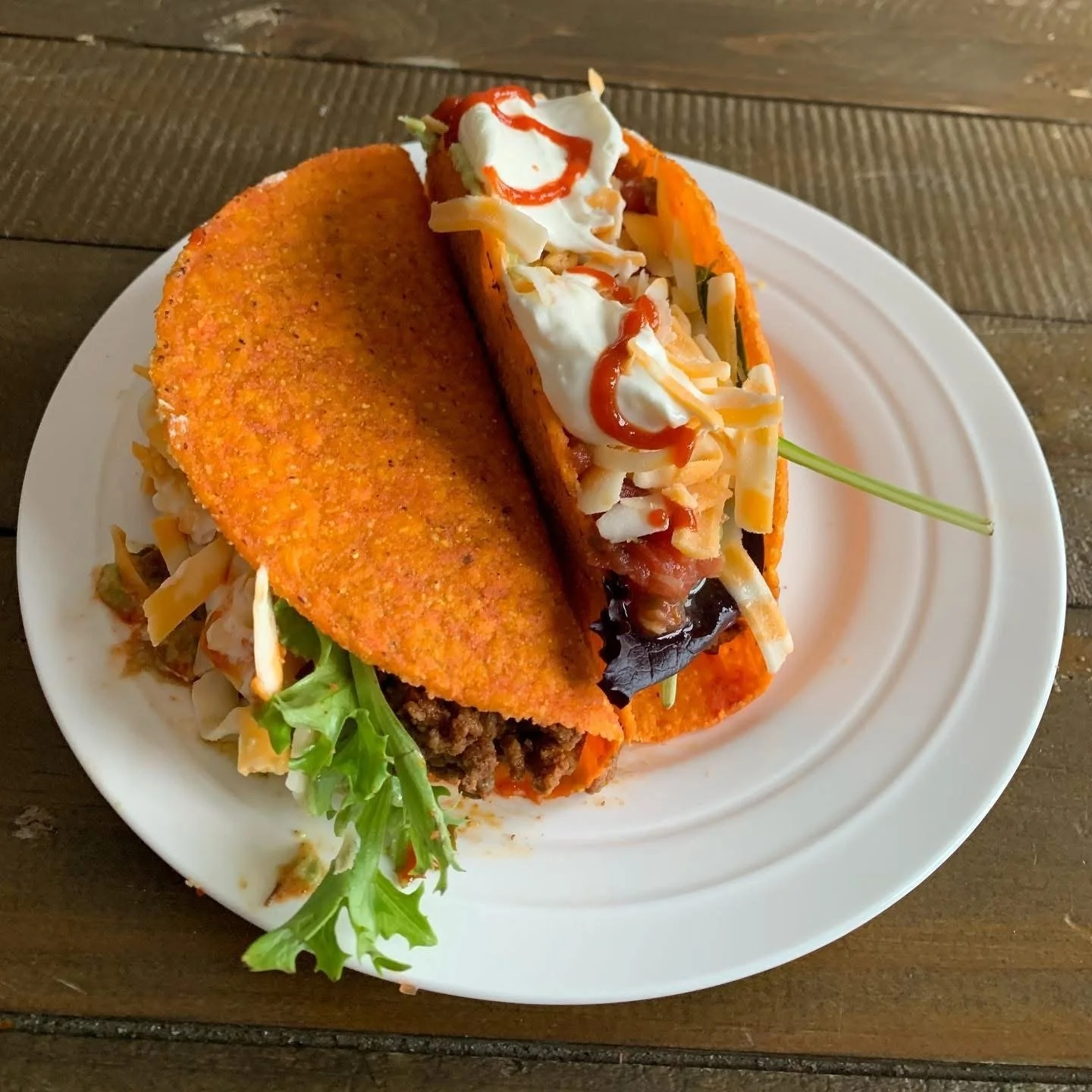 Baja style taco with crispy orange shell, ground beef, shredded cheese, lettuce, sour cream, and hot sauce on a white plate.