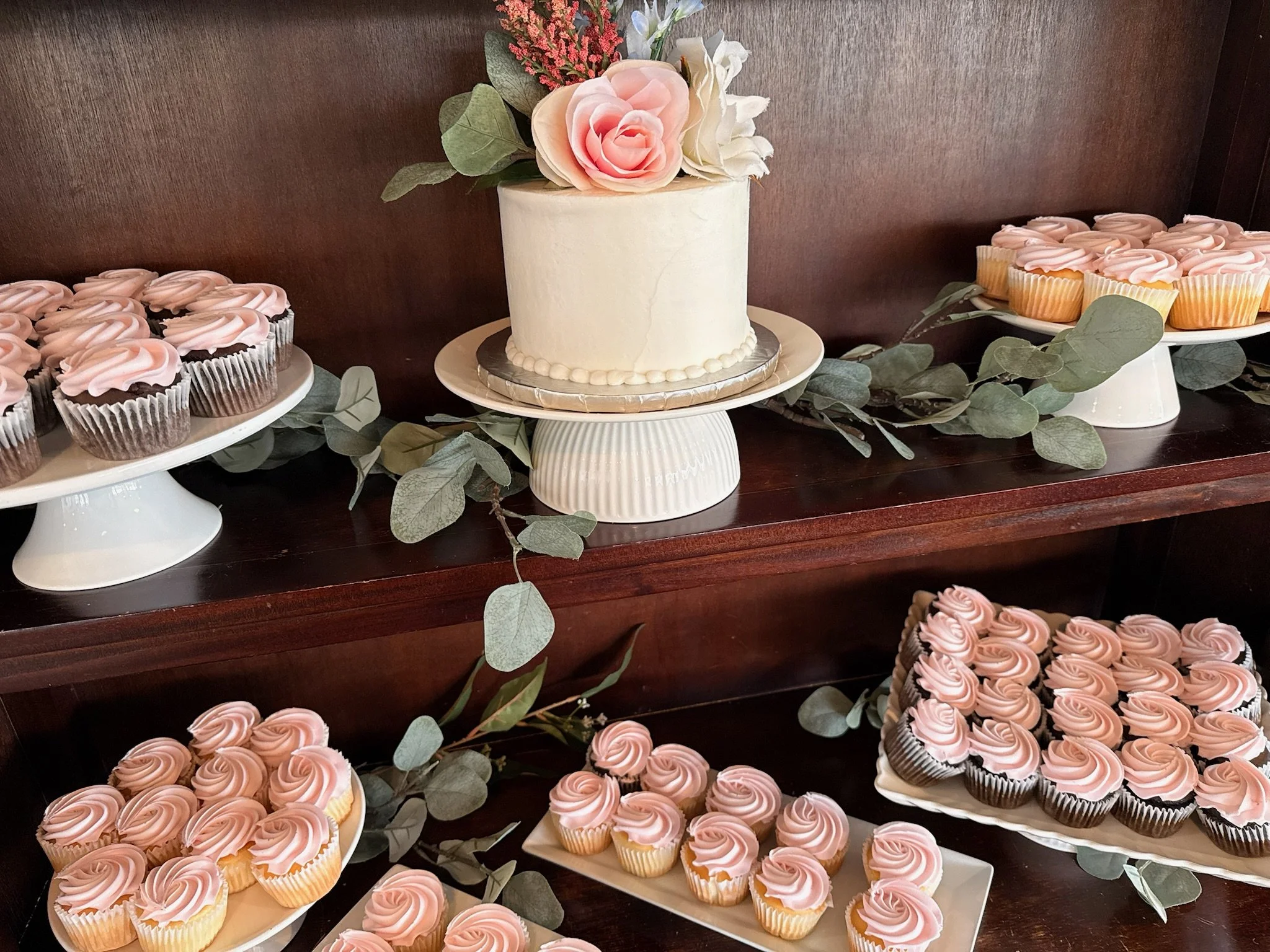 A white cake decorated with pink roses and greenery, surrounded by cupcakes with pink frosting, on wooden shelves with decorative greenery.