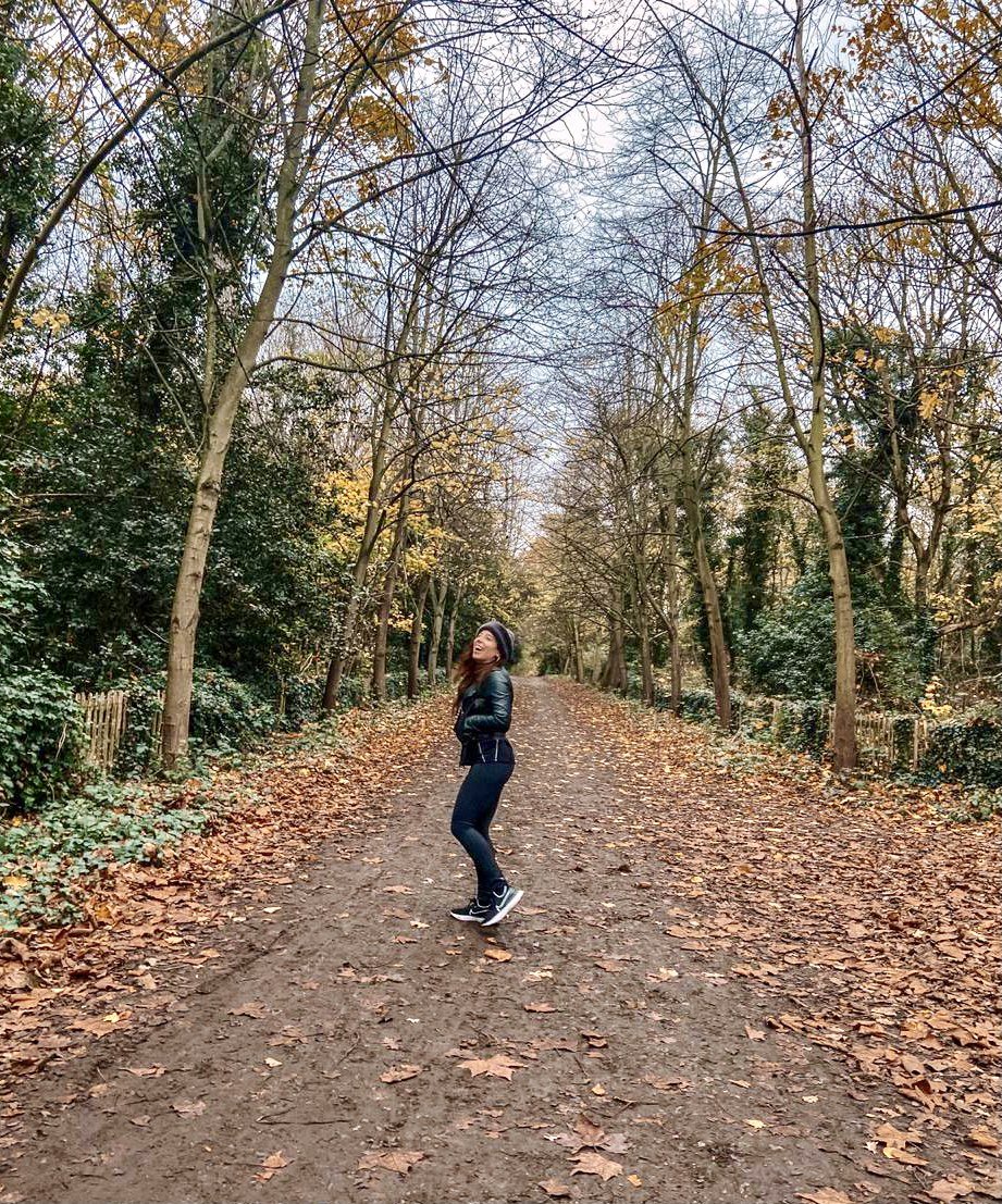 A woman in black athletic clothing and a beanie is walking on a dirt trail in an autumnal forest with fallen leaves.