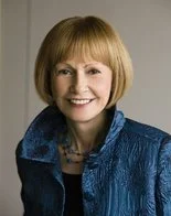 Portrait of a woman with short blonde hair, smiling, wearing a blue jacket and necklace.
