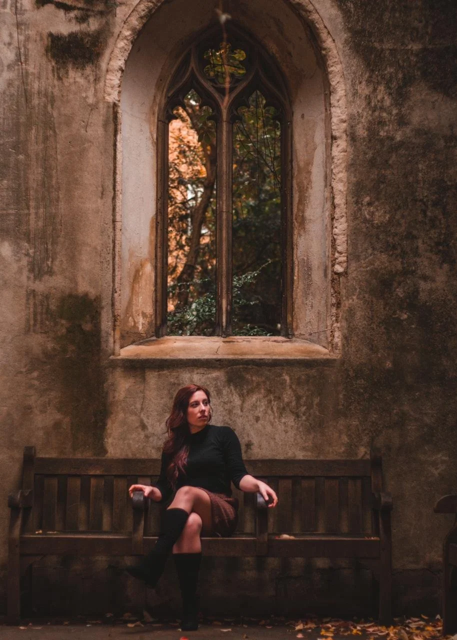 A young woman with long brown hair sitting on a wooden bench inside an old building with a large arched stained glass window behind her. The scene is dimly lit with a view of trees outside the window.