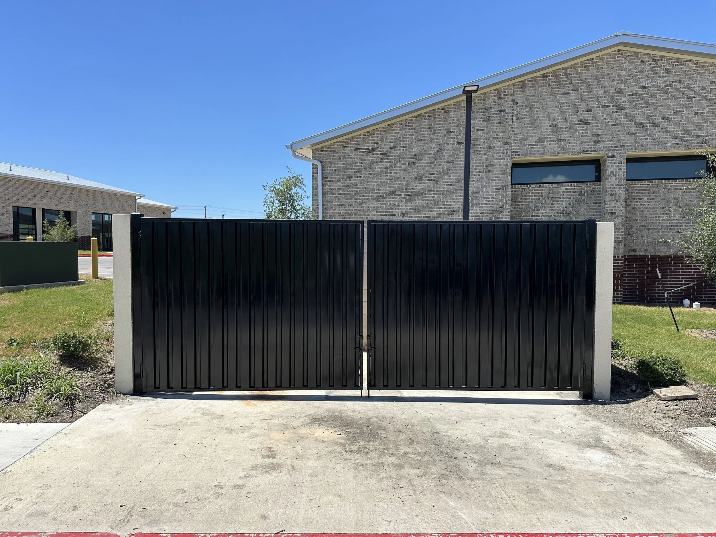 A black metal gate with vertical slats in front of a brick building with rectangular windows under a clear blue sky.