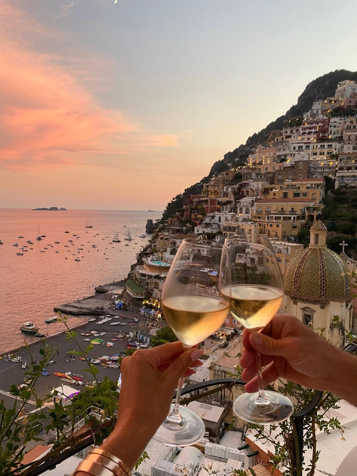 Two people clink glasses of white wine with an Italian coastline and colorful hillside houses in the background during sunset.