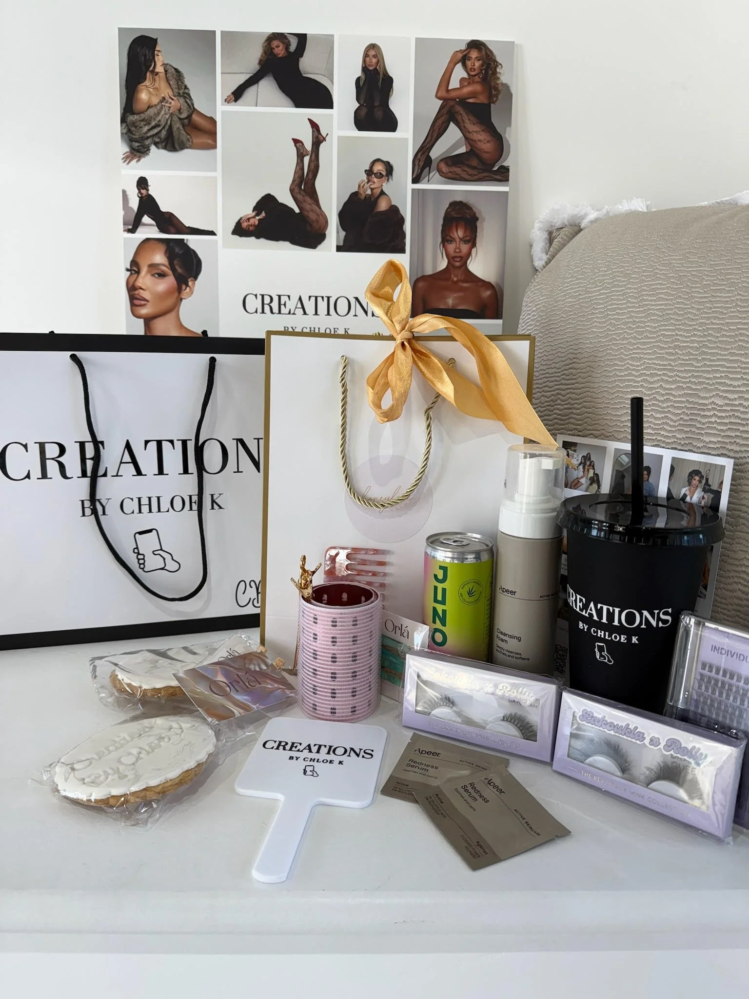 Collection of beauty products and accessories, including a pink eyelash case, ginger cookies, a mirror, skincare bottles, a can of soda, a black cup with a straw, gift bags, and promotional flyers, arranged on a white surface with a poster of model photos in the background.