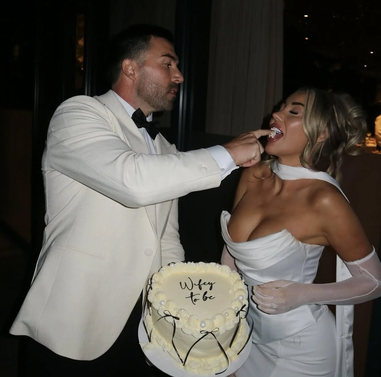 A man in a white tuxedo and a woman in a white dress are at a wedding reception. The man is playfully touching the woman's tongue, which is extending towards his finger. There is a wedding cake with the words 'Wifey to be' on it in front of them.