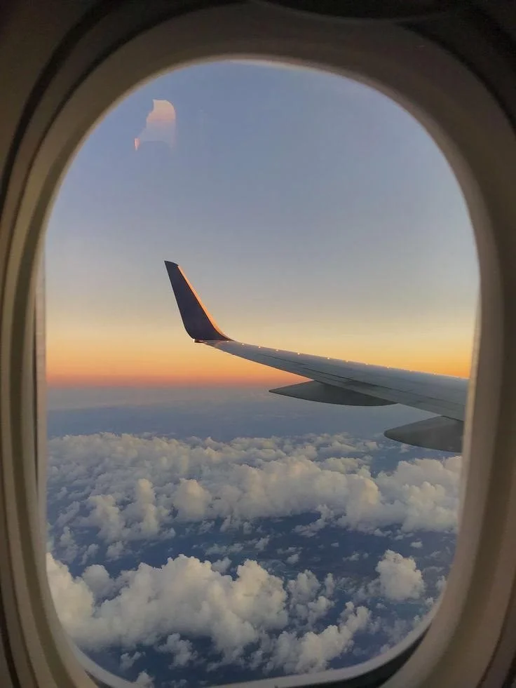 View from an airplane window showing the wing and a sunset sky with clouds below.