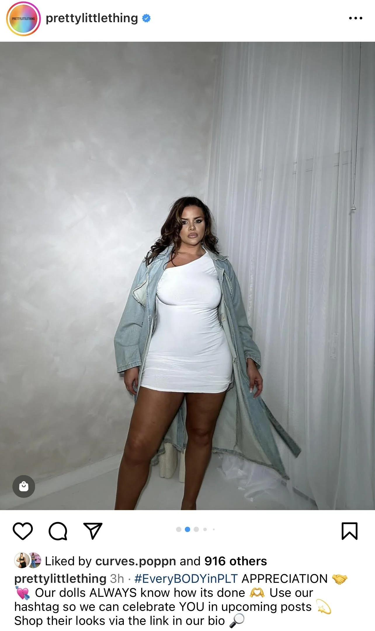 A woman in a white dress and oversized jacket standing against a white wall and sheer curtain.