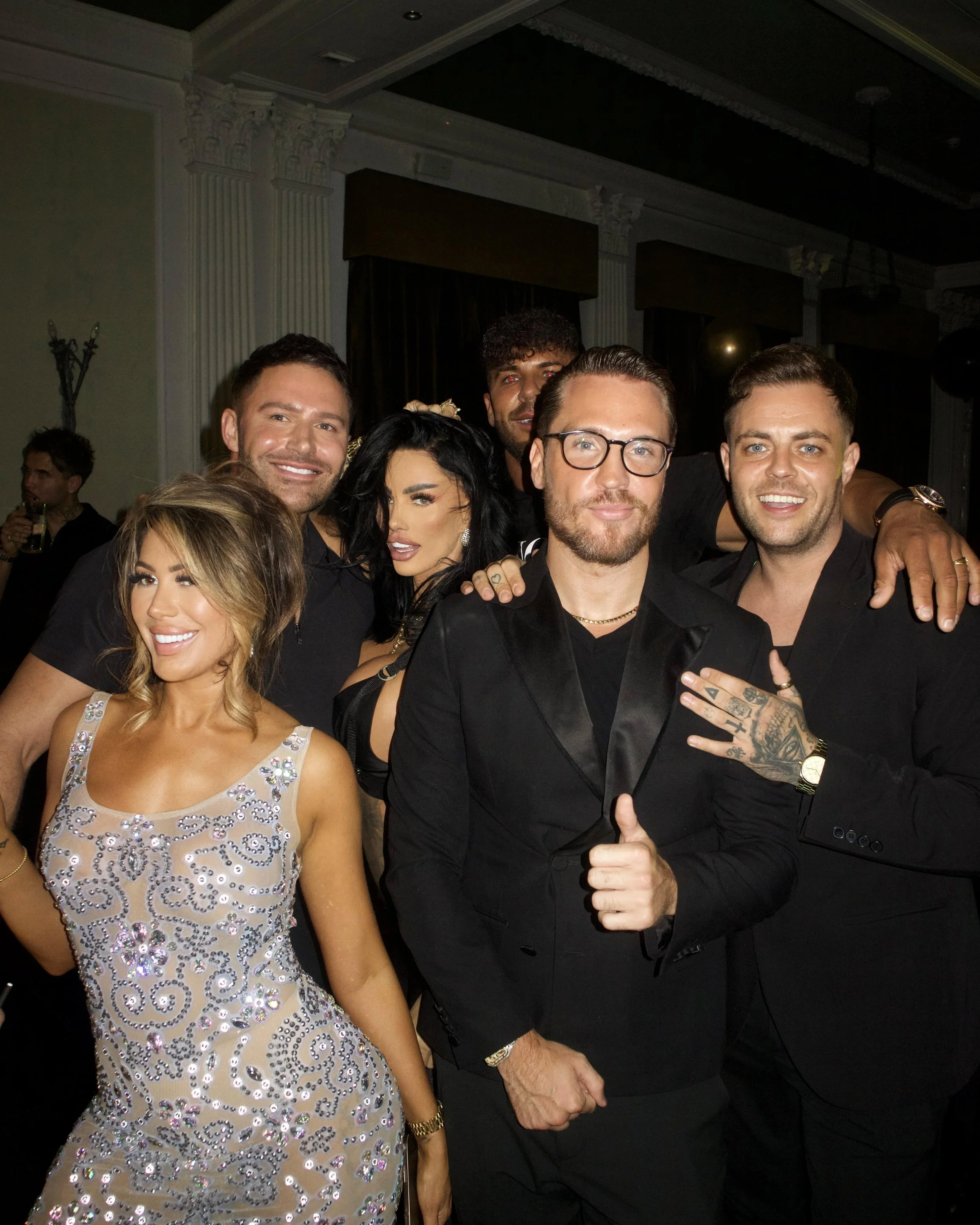Group of seven people posing together at a party or celebration, all dressed in formal or stylish attire, with some smiling and one person giving a thumbs-up.