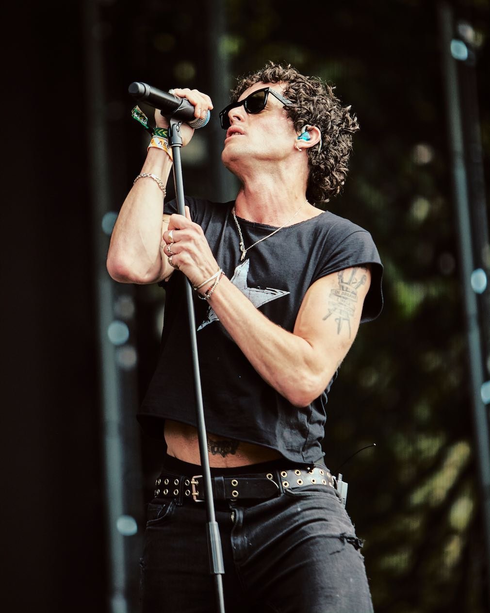 A female singer with curly hair, sunglasses, and tattoos performing on stage with a microphone.