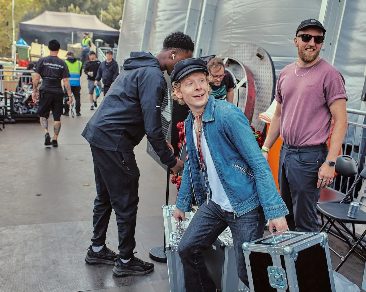 A group of people at an outdoor event, with one person holding a metal case and another person wearing a denim jacket and smiling, while others are in the background near equipment and tents.