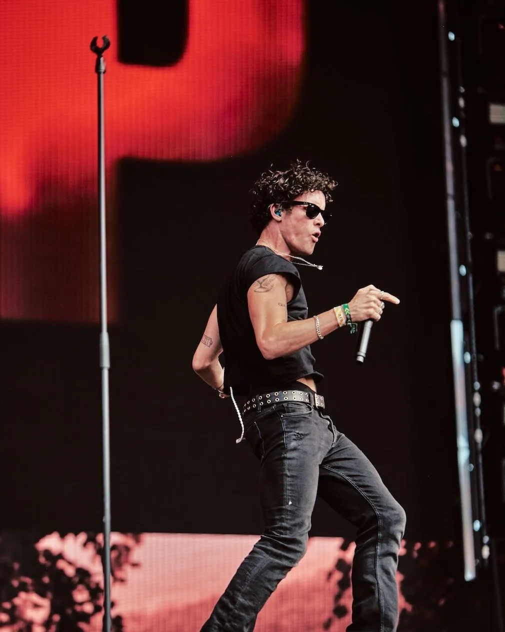 A male singer with curly hair, sunglasses, and tattoos performs on stage holding a microphone, wearing a black sleeveless shirt, black pants, and a studded belt.