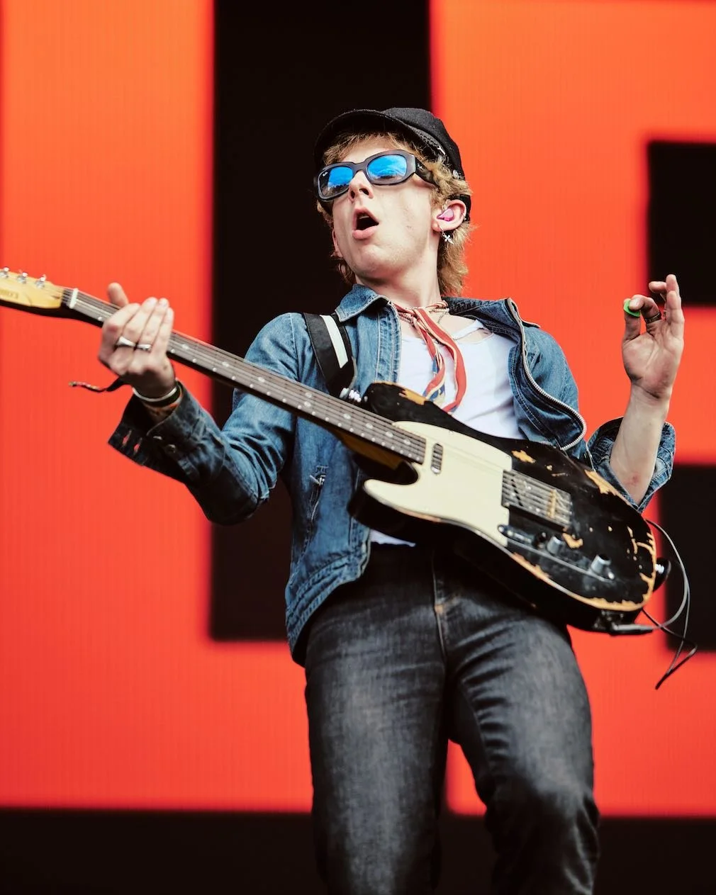 A young man standing on stage playing an electric guitar, wearing sunglasses, a cap, a denim jacket, and a striped scarf, against a red background.