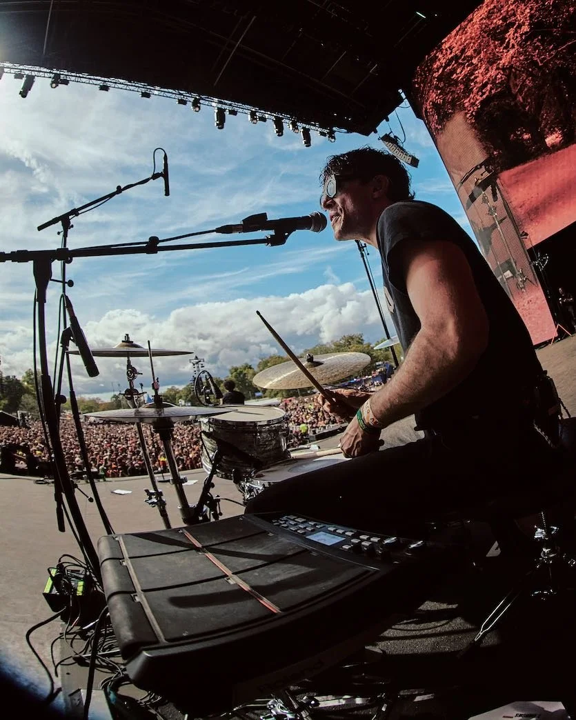 Musician playing electronic drums on stage during an outdoor concert with a large audience and cloudy sky in the background.