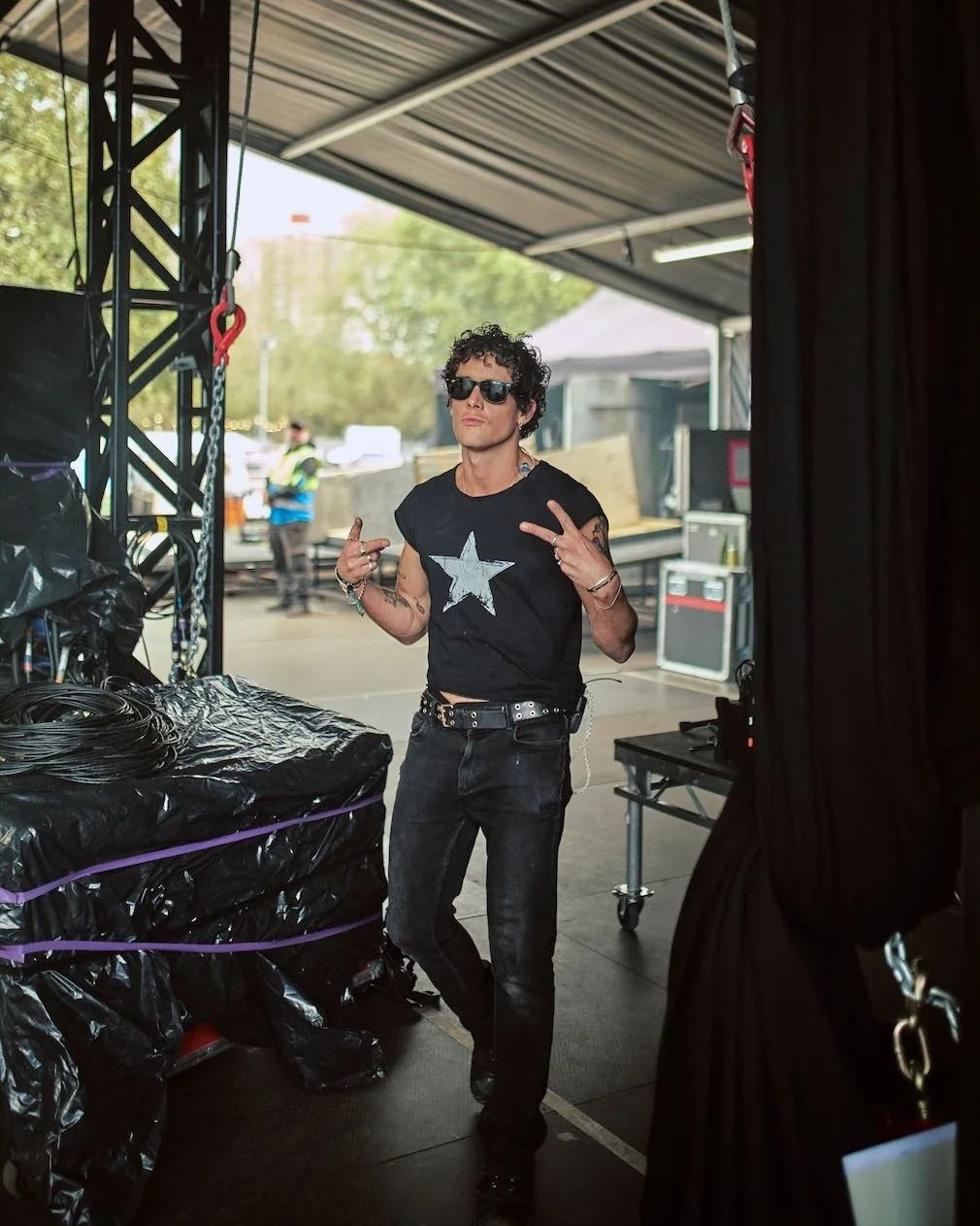 Young man with curly hair, sunglasses, black sleeveless shirt with a star, and black jeans, posing backstage behind stage equipment, making peace signs with his fingers.