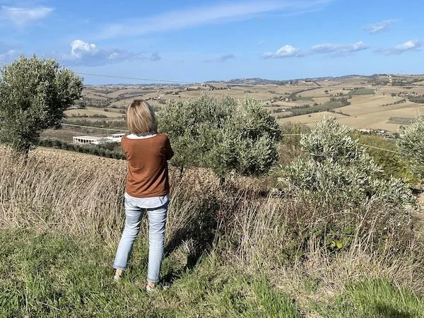 Claudia Wiebe standing on grassy land, and taking a picture of the scenic valley with rolling hills, olive trees, and clear blue sky outside Fano, Italy.
