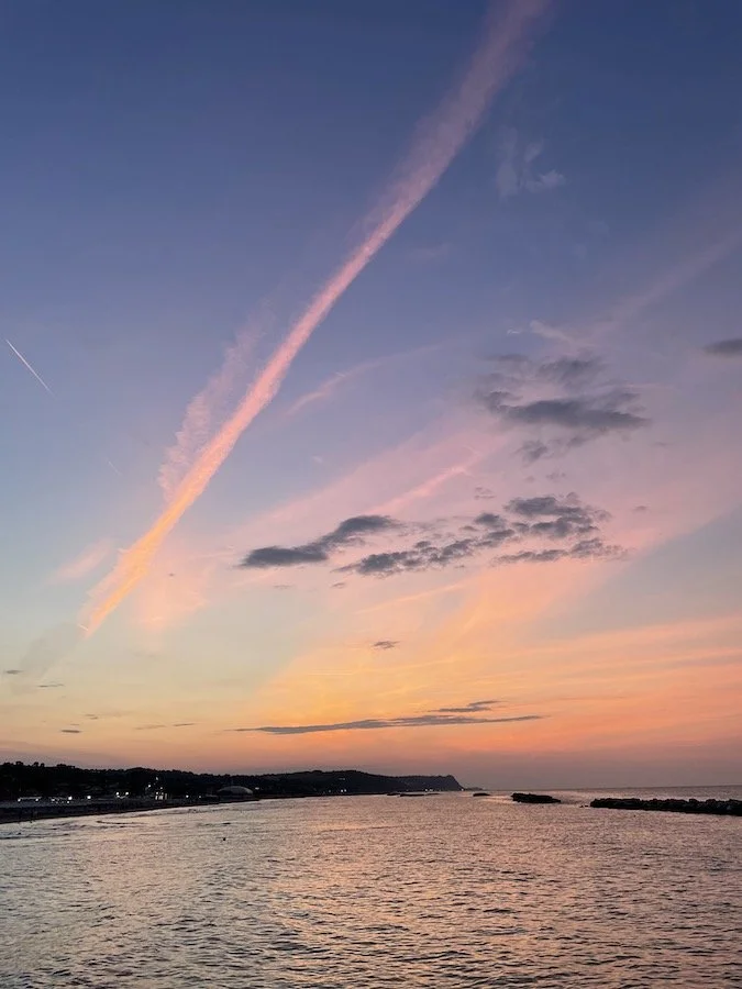 Sunset in Fano, Italy, over a calm ocean with pink and orange streaks in the sky, reflecting on the water, with a distant coastline visible.