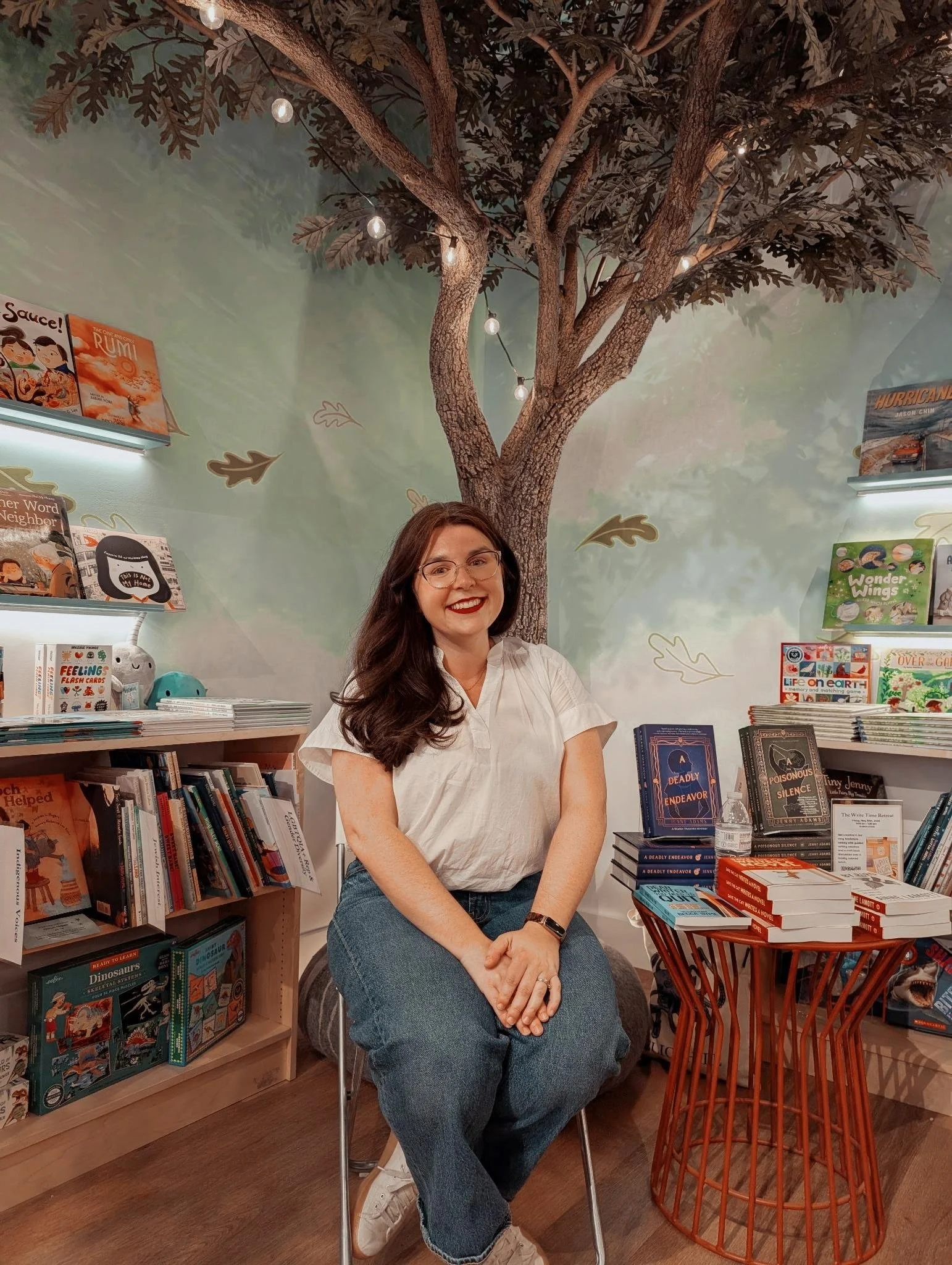A woman with long dark hair, glasses, and a white shirt sitting on a chair inside a bookstore with bookshelves, a mural of a tree on the wall, and fairy lights hanging from the ceiling.