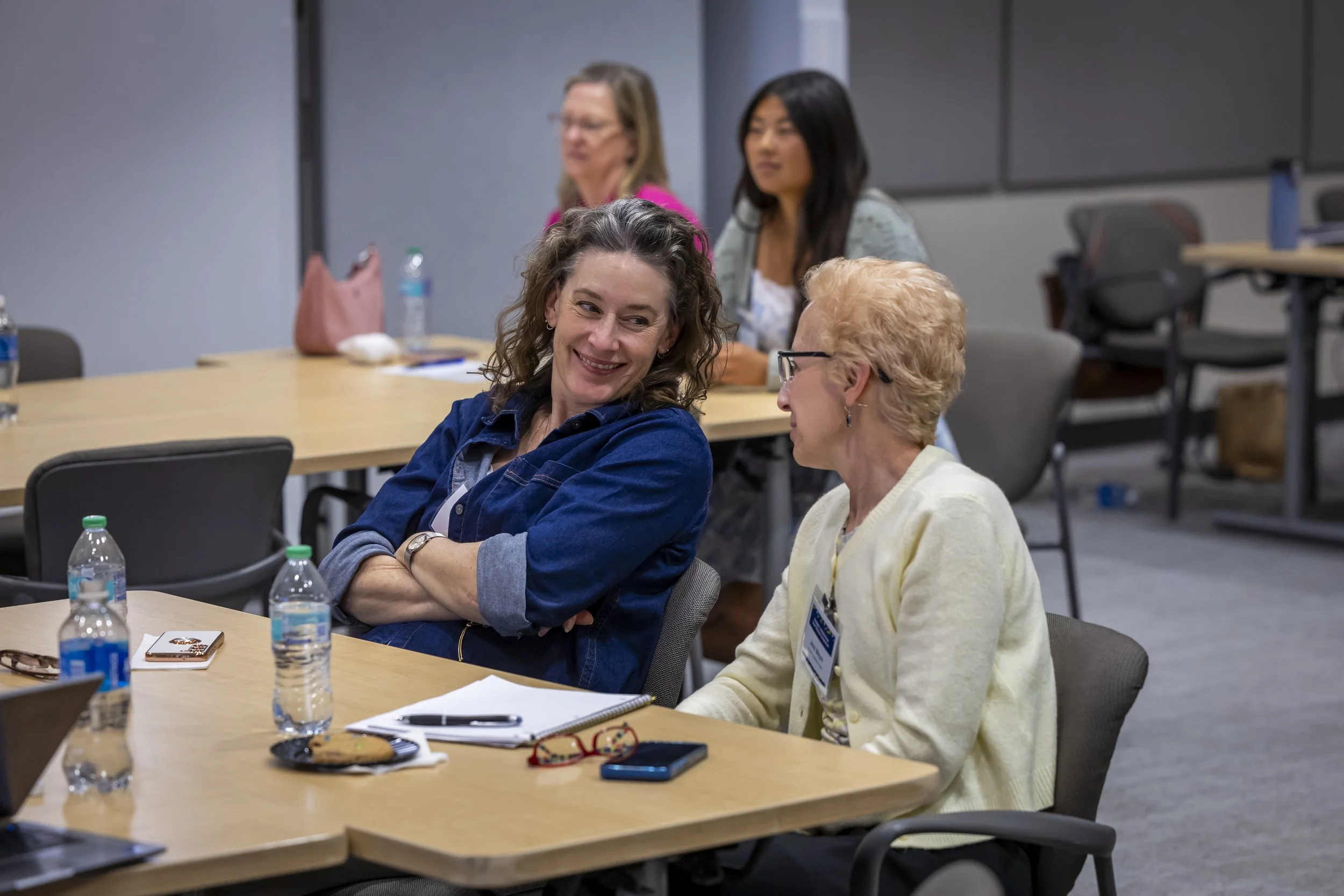 A group of women seated around a conference table engaging in conversation, with notebooks, water bottles, and snacks on the table in a professional setting.