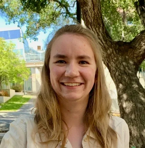 Young woman smiling outdoors in front of a tree with green leaves, buildings, and a blue sky in the background.