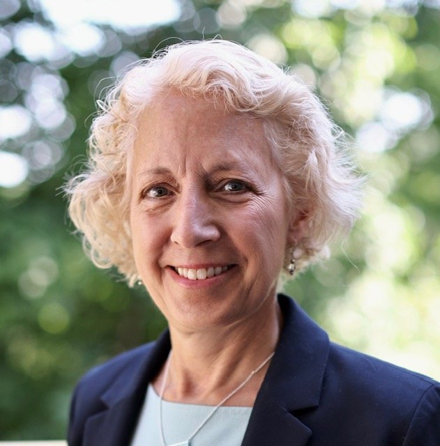 A smiling elderly woman with curly blonde hair, wearing a navy blue blazer and a white top, standing outdoors with blurred green trees in the background.