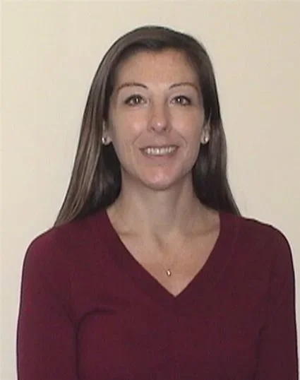 A woman with long brown hair wearing a burgundy top, smiling, standing against a plain light-colored background.