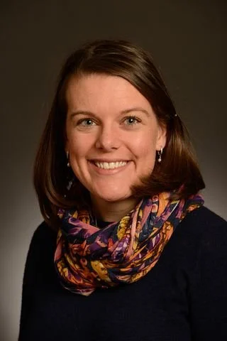 A smiling woman with shoulder-length brown hair, wearing a colorful scarf and earrings, against a plain dark background.