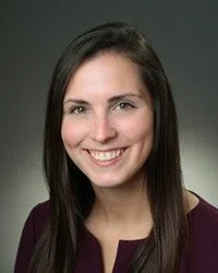 A woman with long brown hair smiling, wearing a dark blazer, against a dark gradient background.