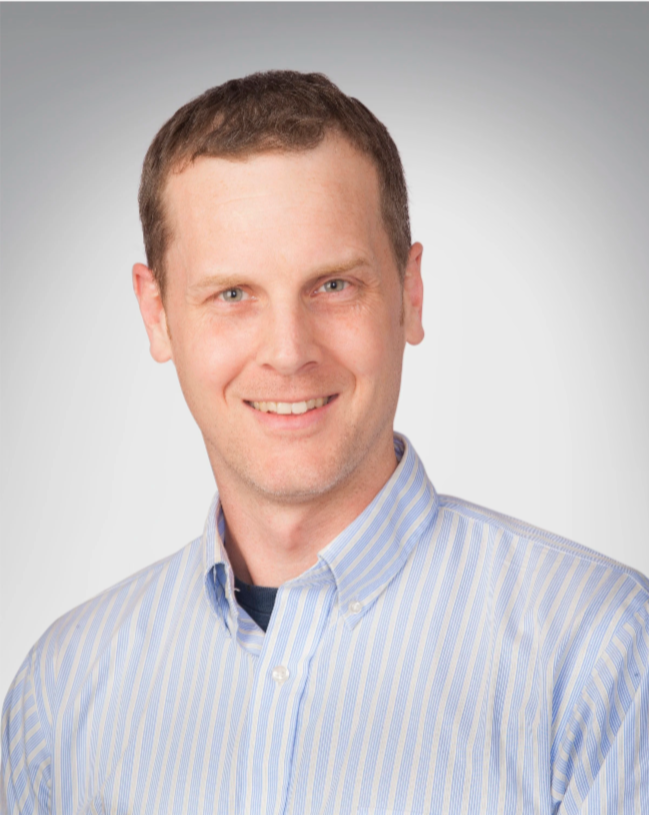 Portrait of a smiling man wearing a light blue striped button-up shirt against a neutral gray background.