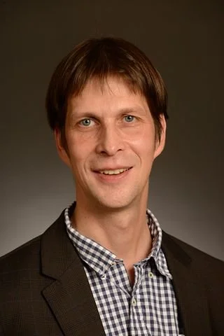 Professional portrait of a man with brown hair, wearing a checkered shirt and a dark blazer, smiling against a plain background.
