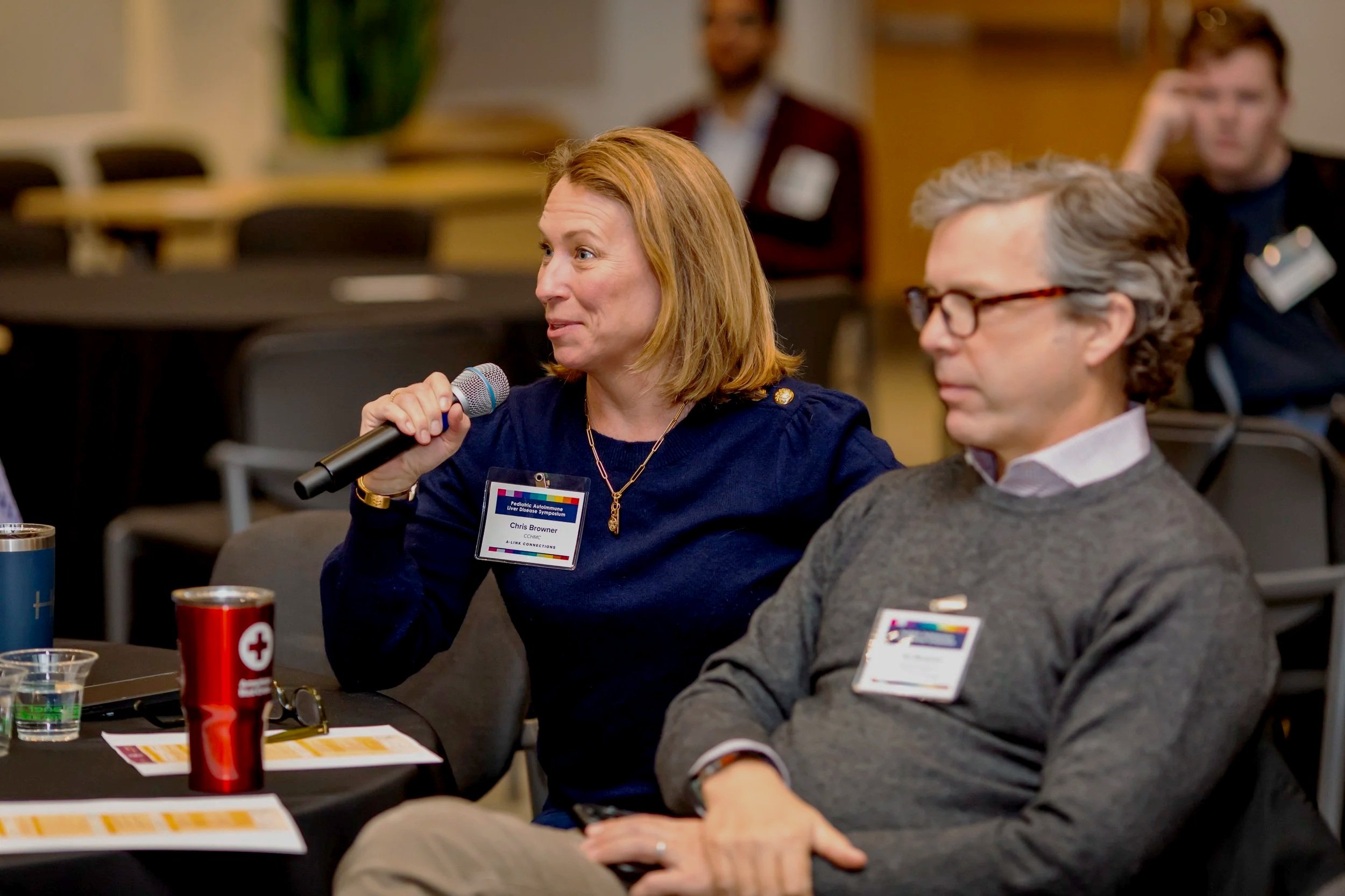 A woman with shoulder-length red hair speaking into a microphone at a conference or workshop, with two men seated next to her listening; one man has gray hair and glasses, and the other has dark hair; table with brochures, coffee mugs, and glasses in front of them.