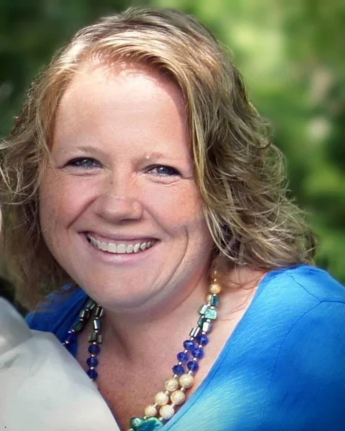 Smiling woman with curly blonde hair wearing a blue top and beaded necklace outdoors.