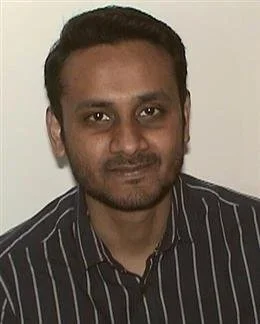 Headshot of a man with short dark hair, wearing a dark striped shirt, against a plain light background.