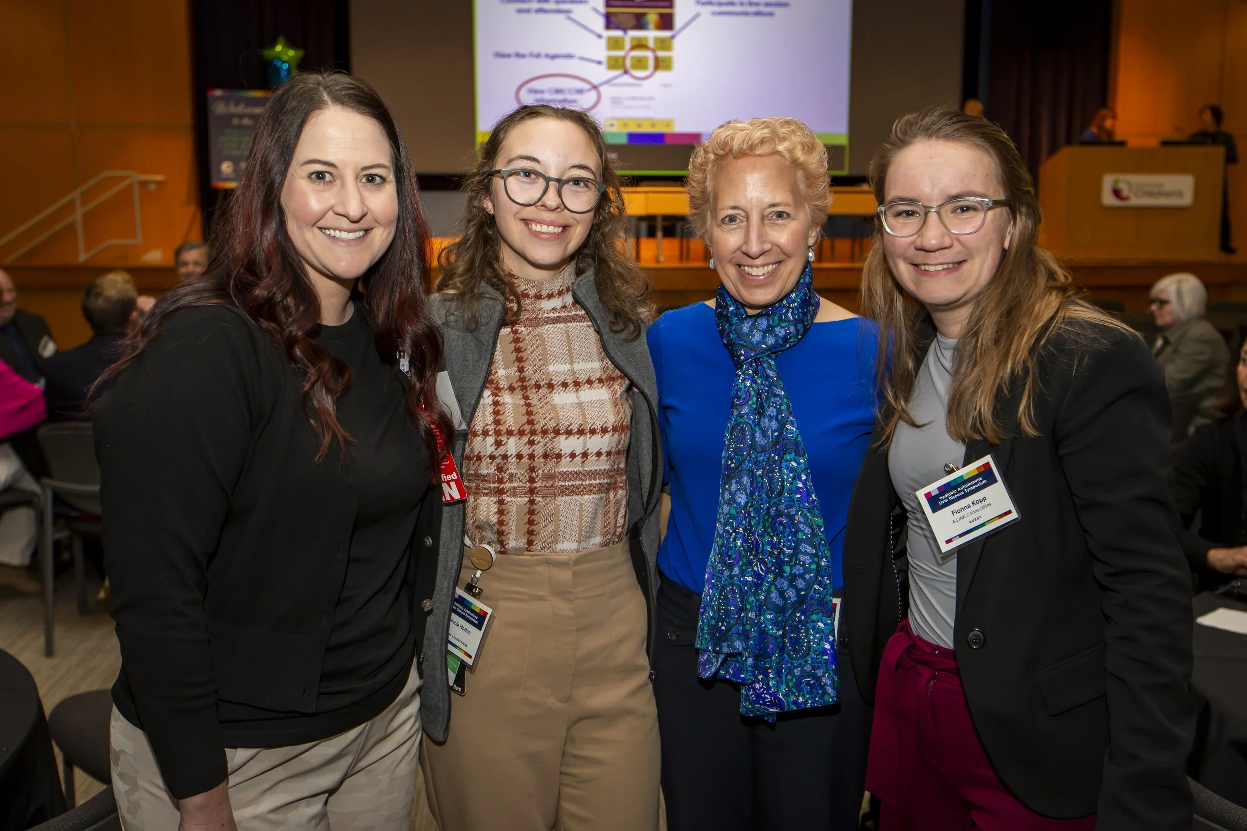 Four women standing close together at a conference, smiling at the camera, with a presentation slide and other attendees in the background.