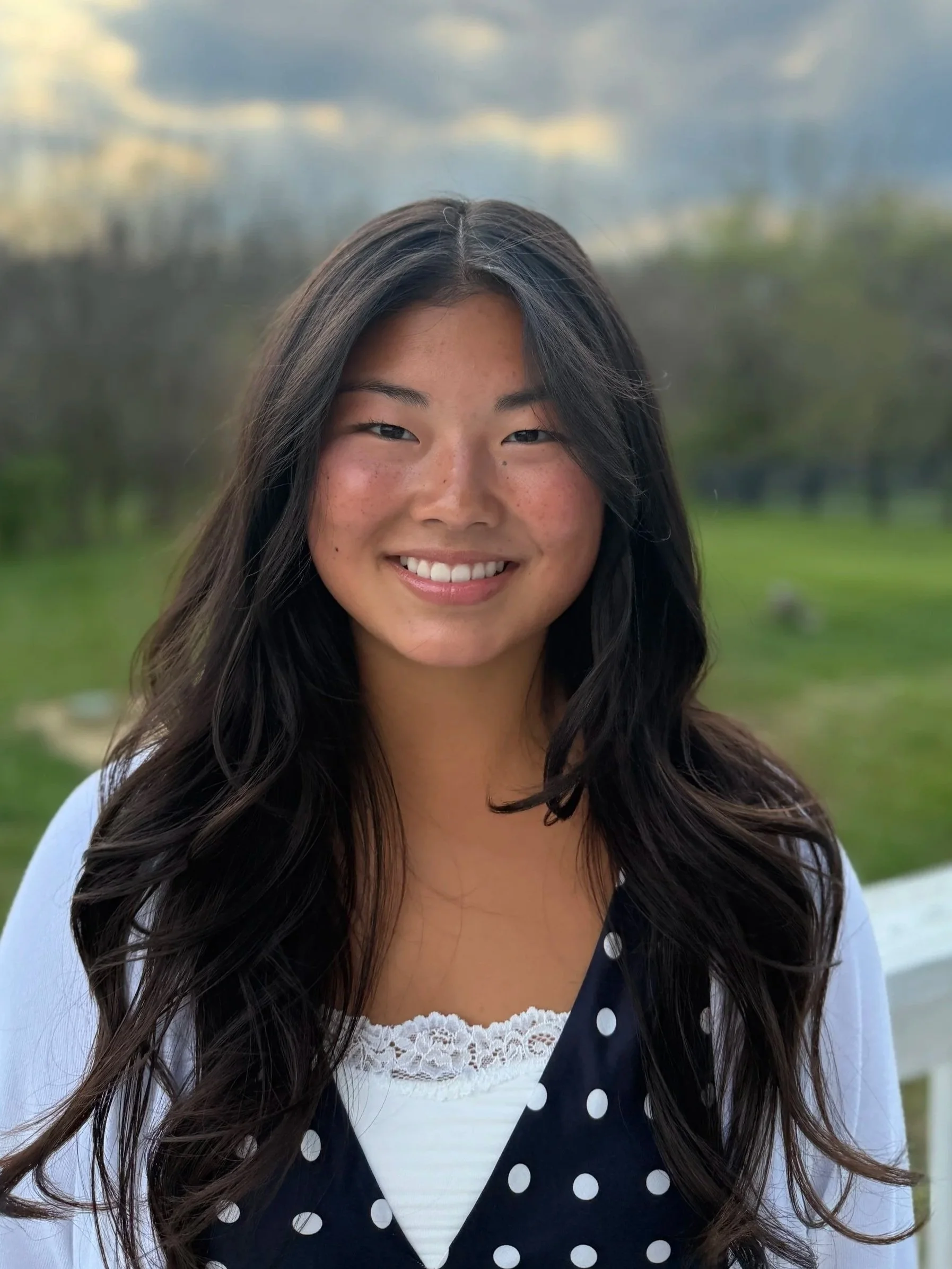 A young woman with long, wavy black hair, tan skin, and freckles, smiling outdoors with a blurred background of trees and cloudy sky.