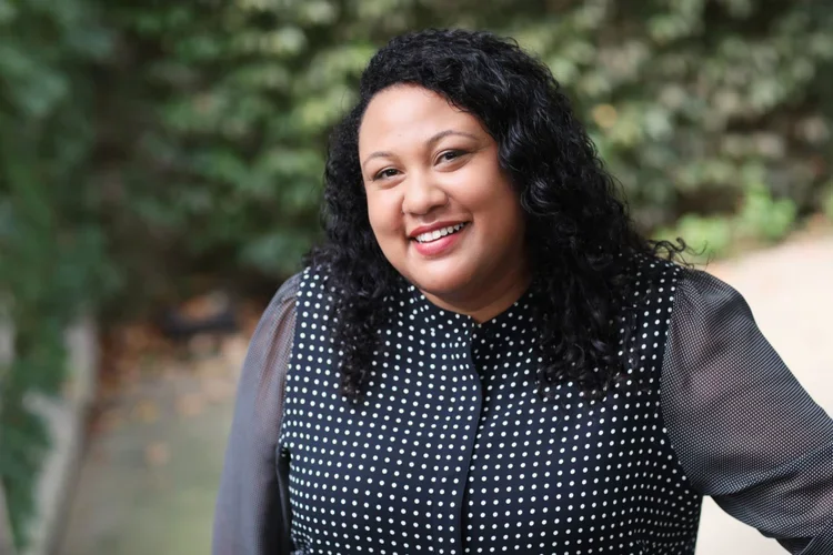A woman with dark curly hair smiling outdoors, wearing a black and white polka dot blouse.
