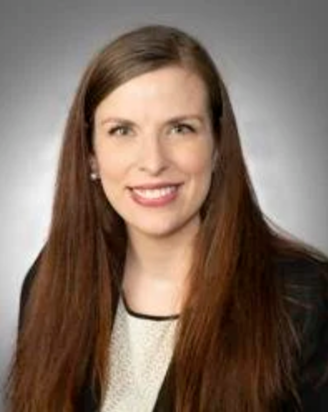 Headshot of a woman with long brown hair, smiling, wearing a black blazer and white top, against a gray background.
